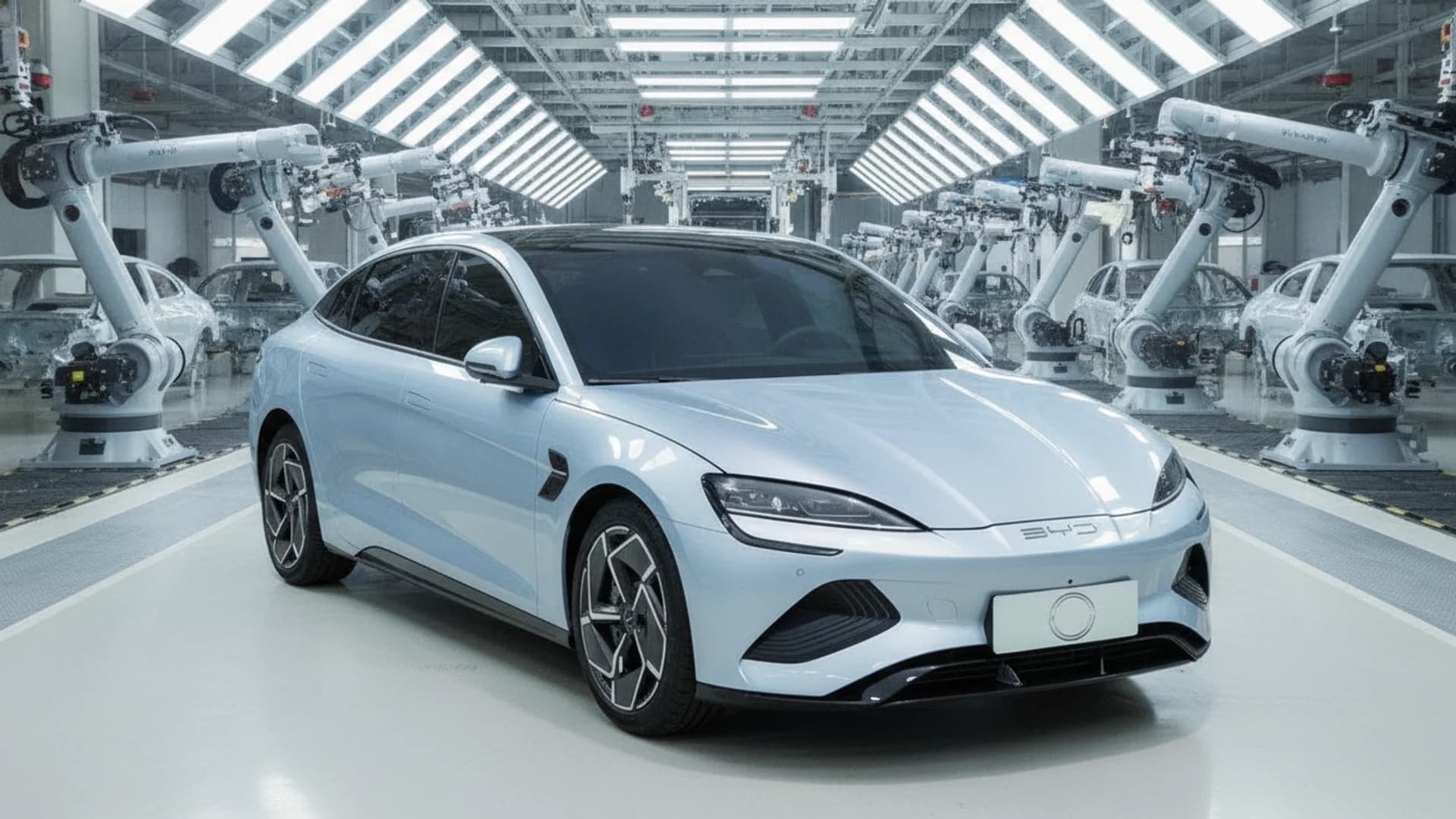 A light blue sedan sits centered on a factory assembly line surrounded by white robotic arms and overhead lights.