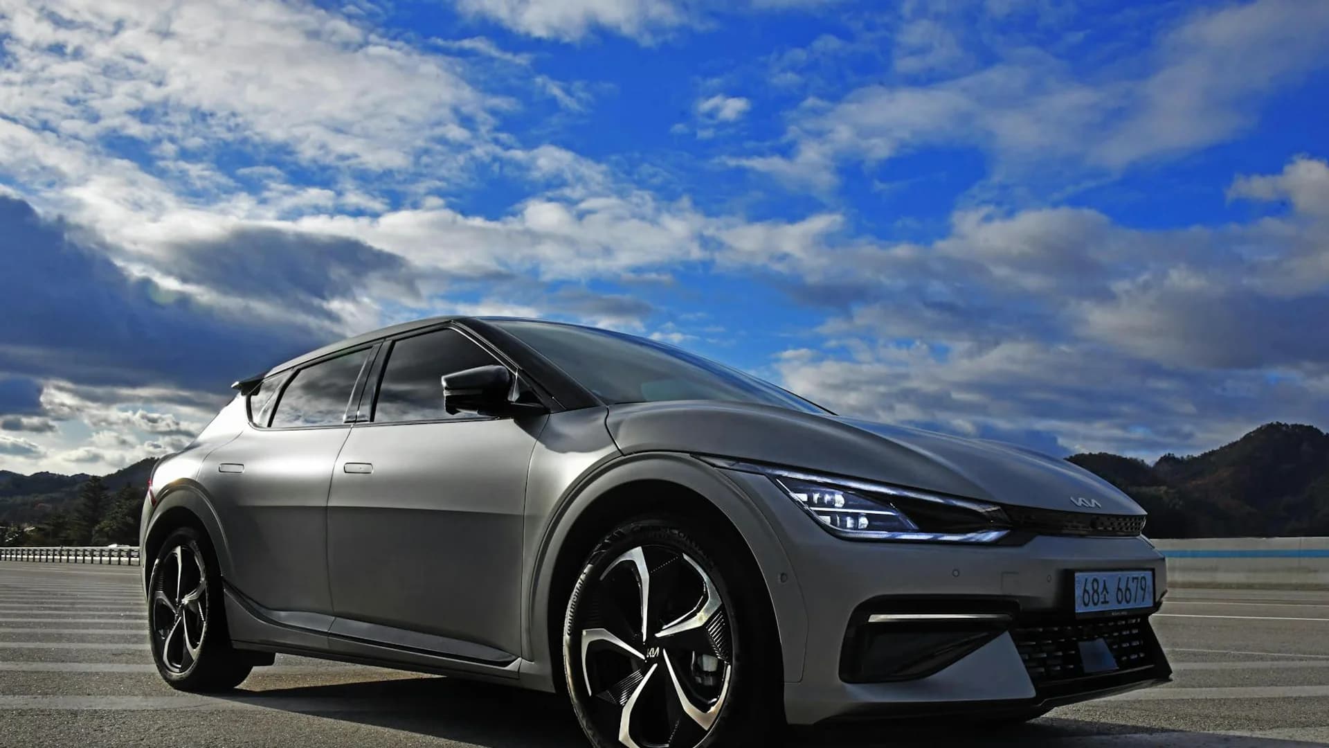 A silver Kia EV6 is parked on an asphalt road under a bright blue sky with white clouds.