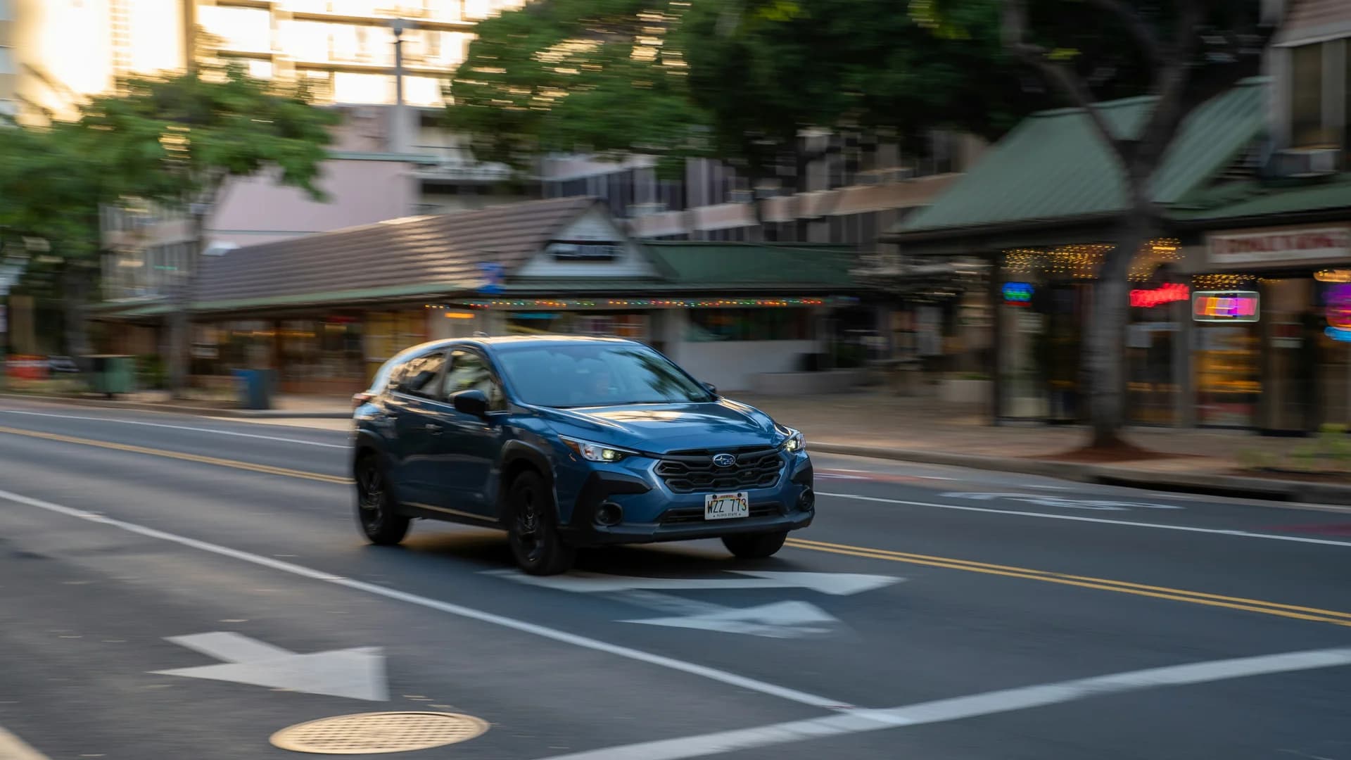 A blue SUV drives on a city street with blurred buildings and trees in the background.