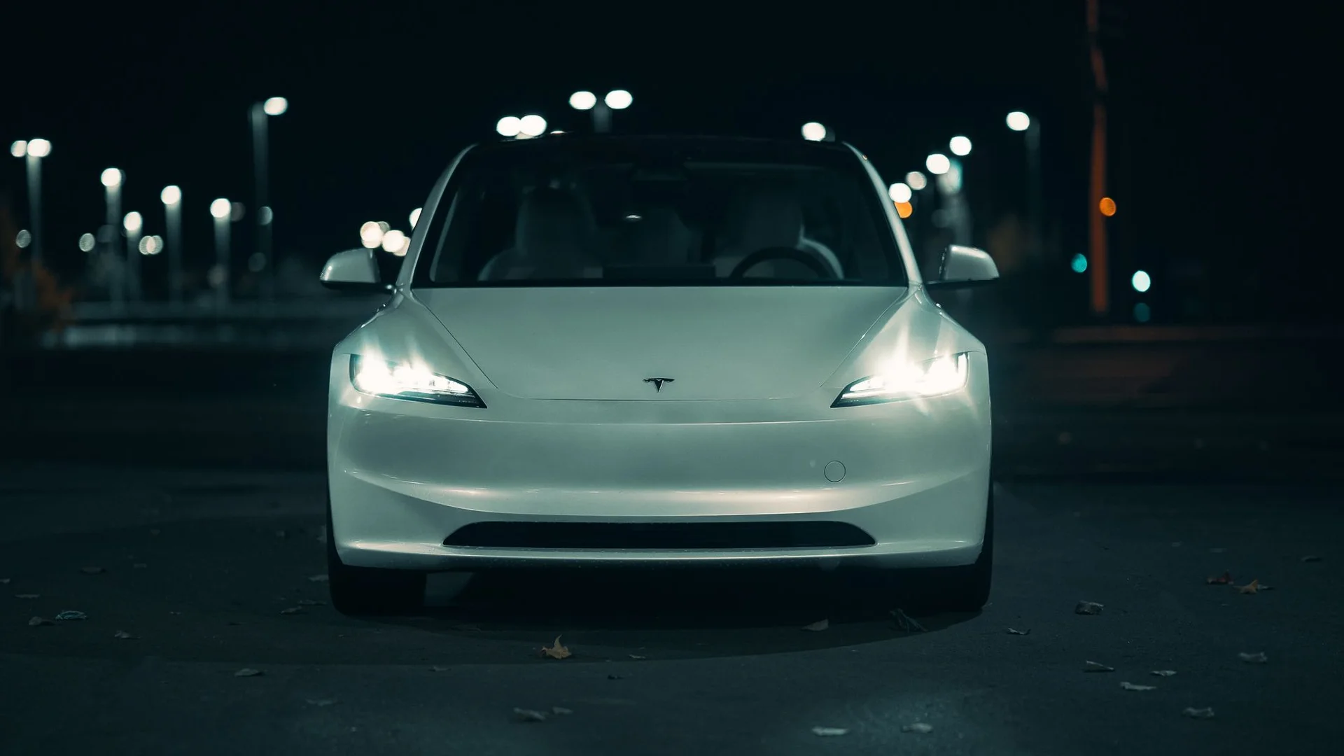 White Tesla car with headlights on, parked at night with blurred streetlights in the background.