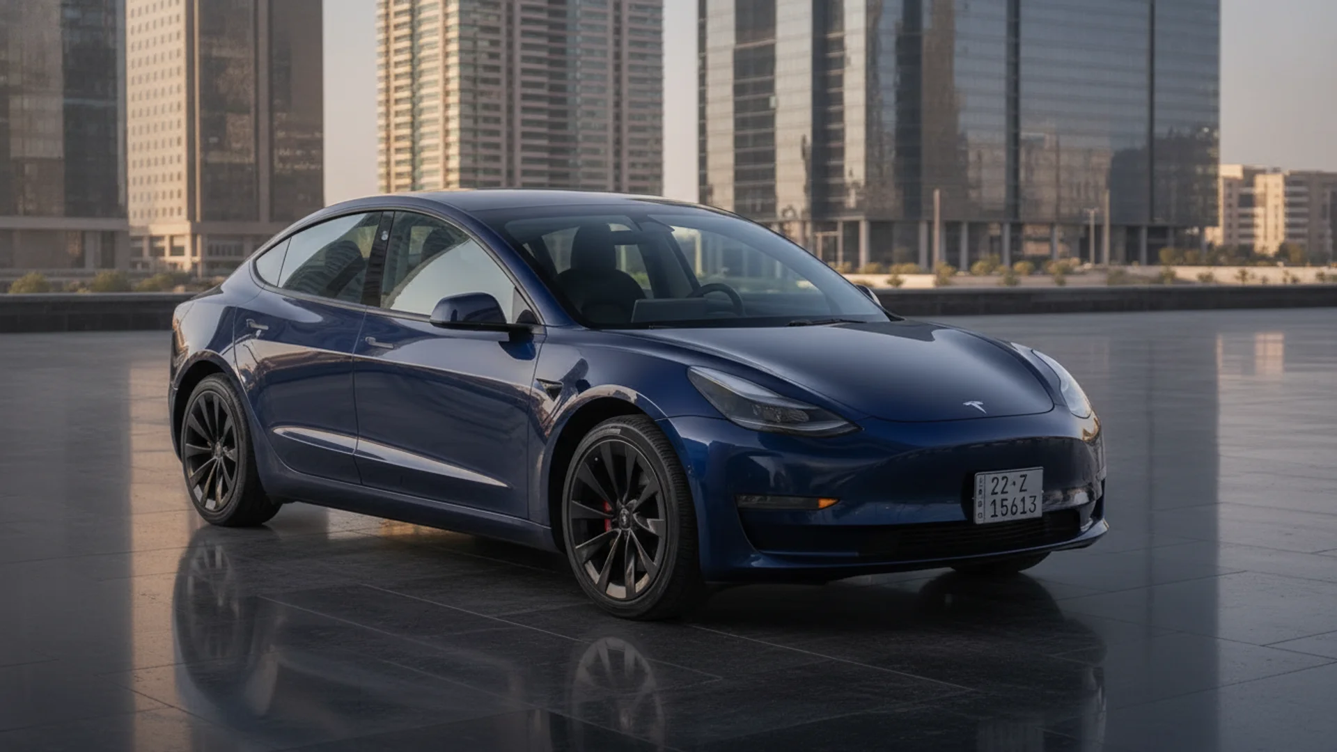 A dark blue Tesla Model 3 is parked on a reflective surface with tall buildings in the background.