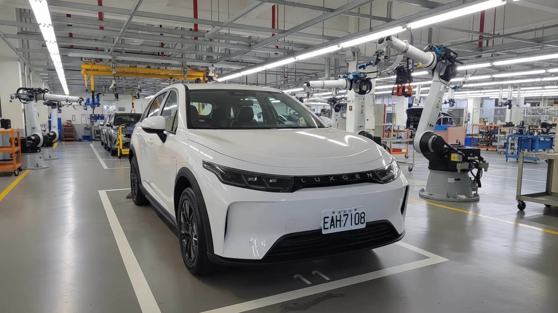 A white Luxgen electric SUV parked inside a high-tech manufacturing facility with robotic assembly arms.