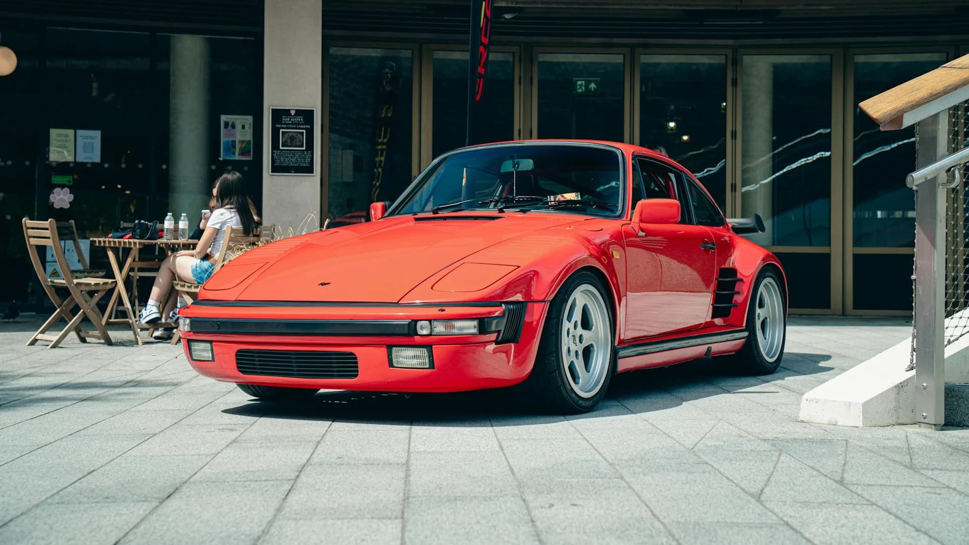 A bright red sports car is parked on a grey tiled sidewalk in front of a building with glass doors.