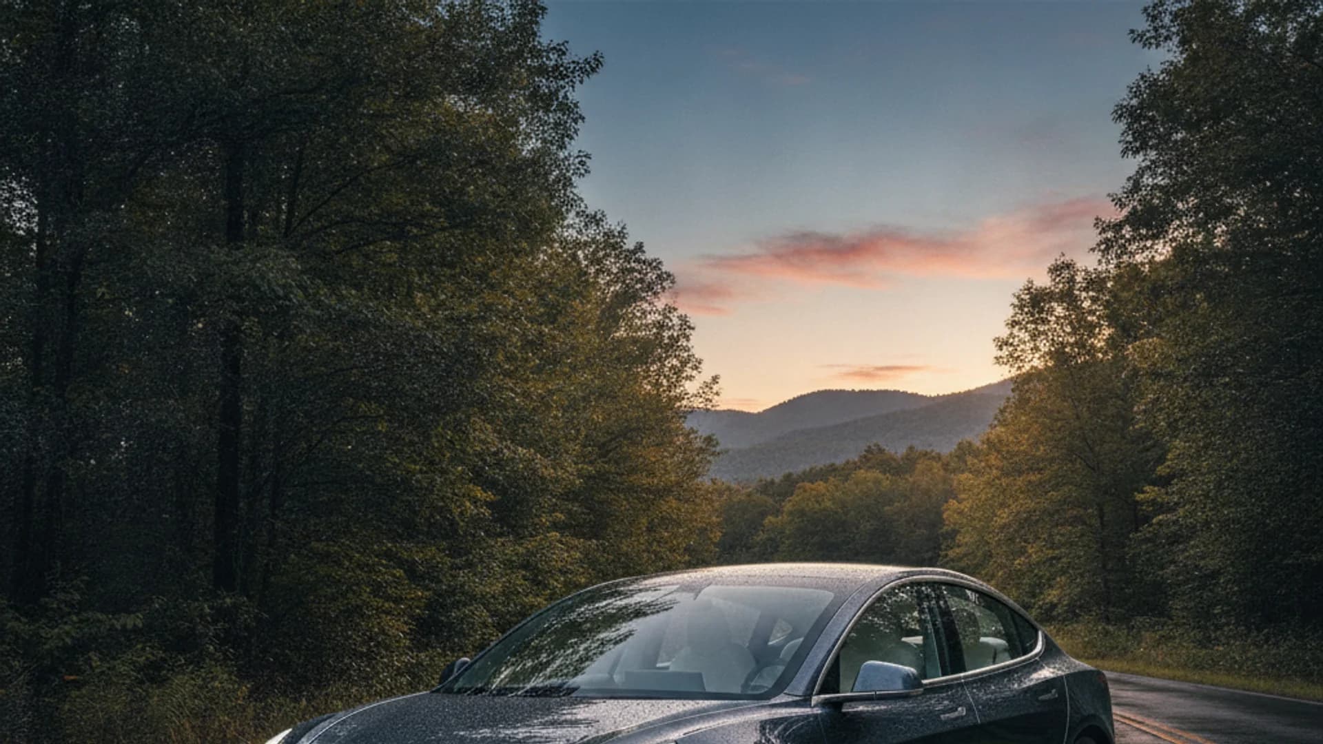 A dark grey car with water droplets on its hood is parked on a road surrounded by trees, with mountains and a sunset sky in the background.