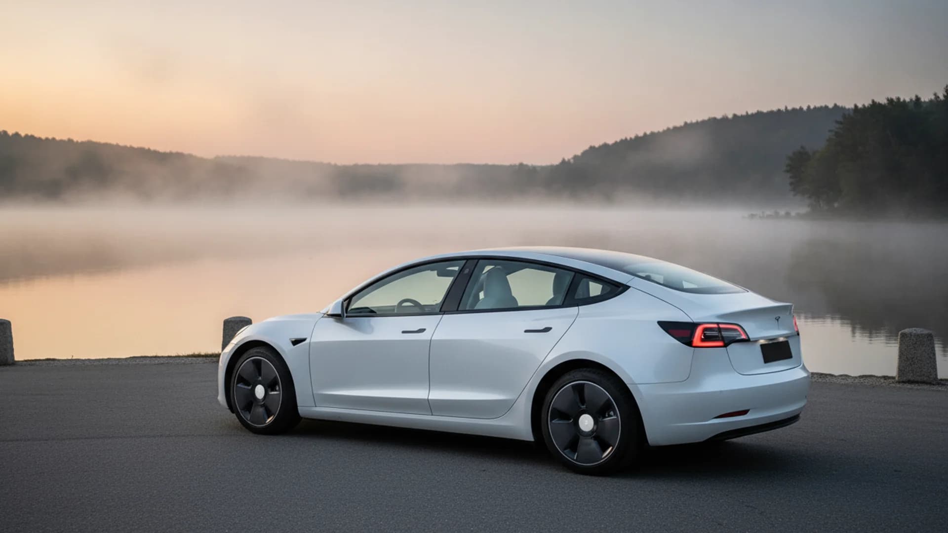 A white Tesla Model 3 is parked on asphalt, facing right, with a foggy lake and trees in the background.