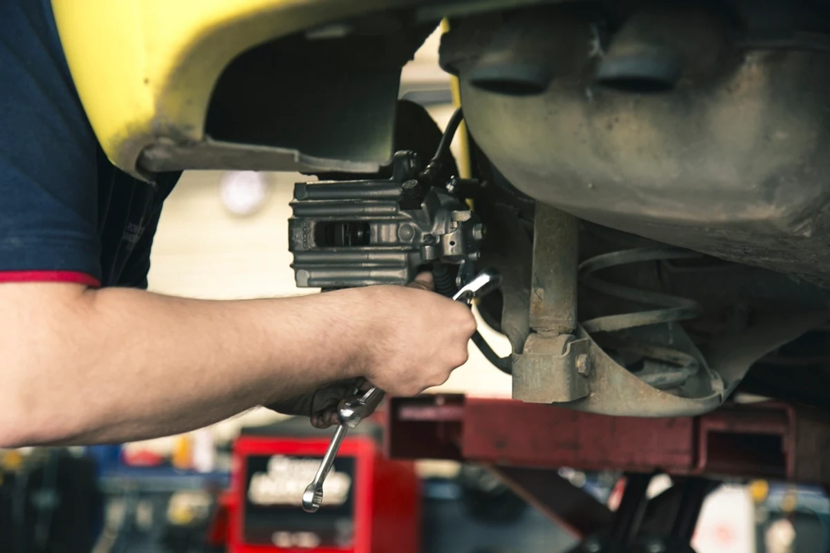 Technician working on a brake caliper and front suspension under a vehicle raised on a shop hoist